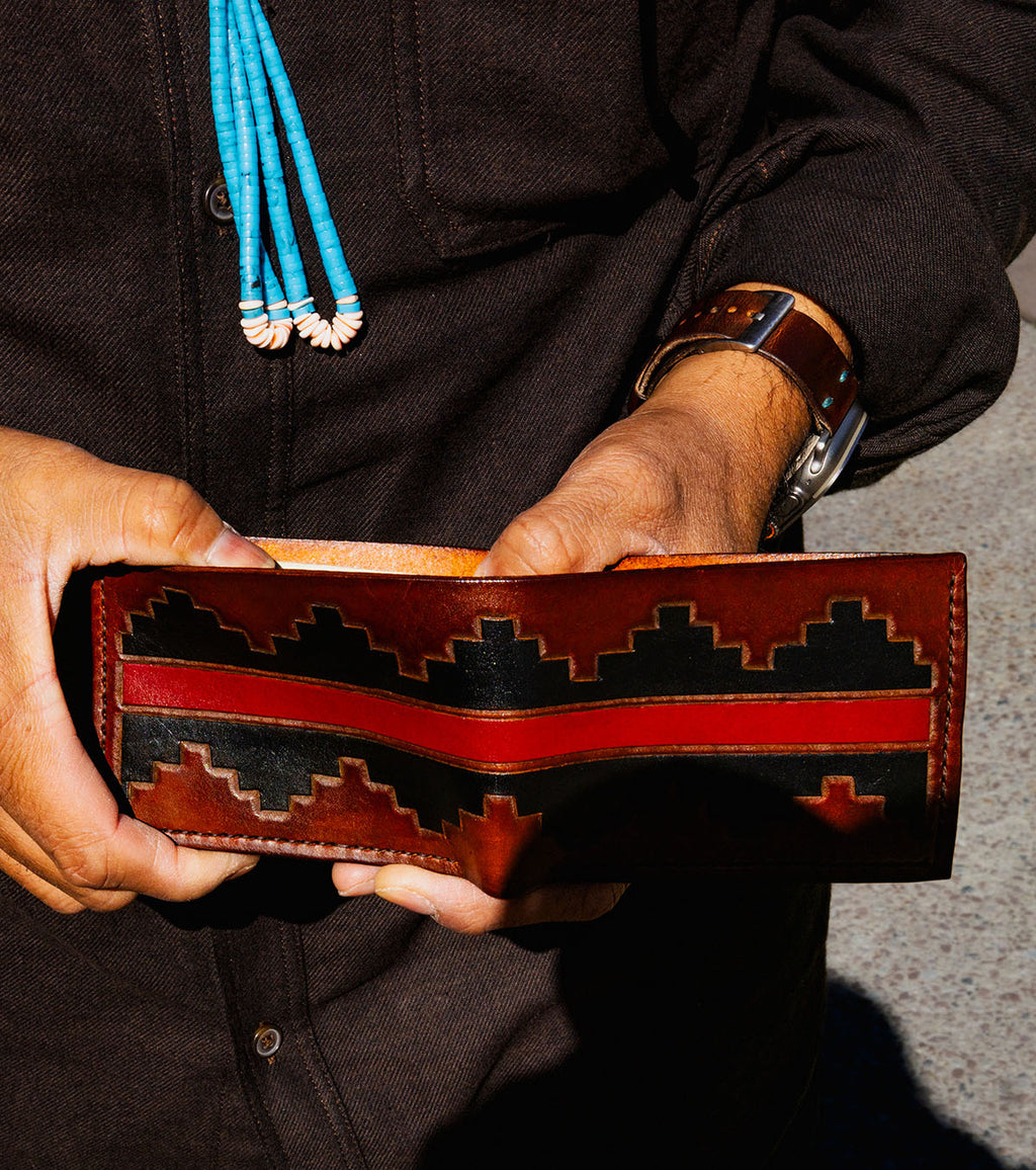 A close-up of a person holding a brown leather bi-fold wallet with a black and red geometric pattern. The person is wearing a dark brown shirt, a turquoise and white beaded necklace, and a brown leather watch.