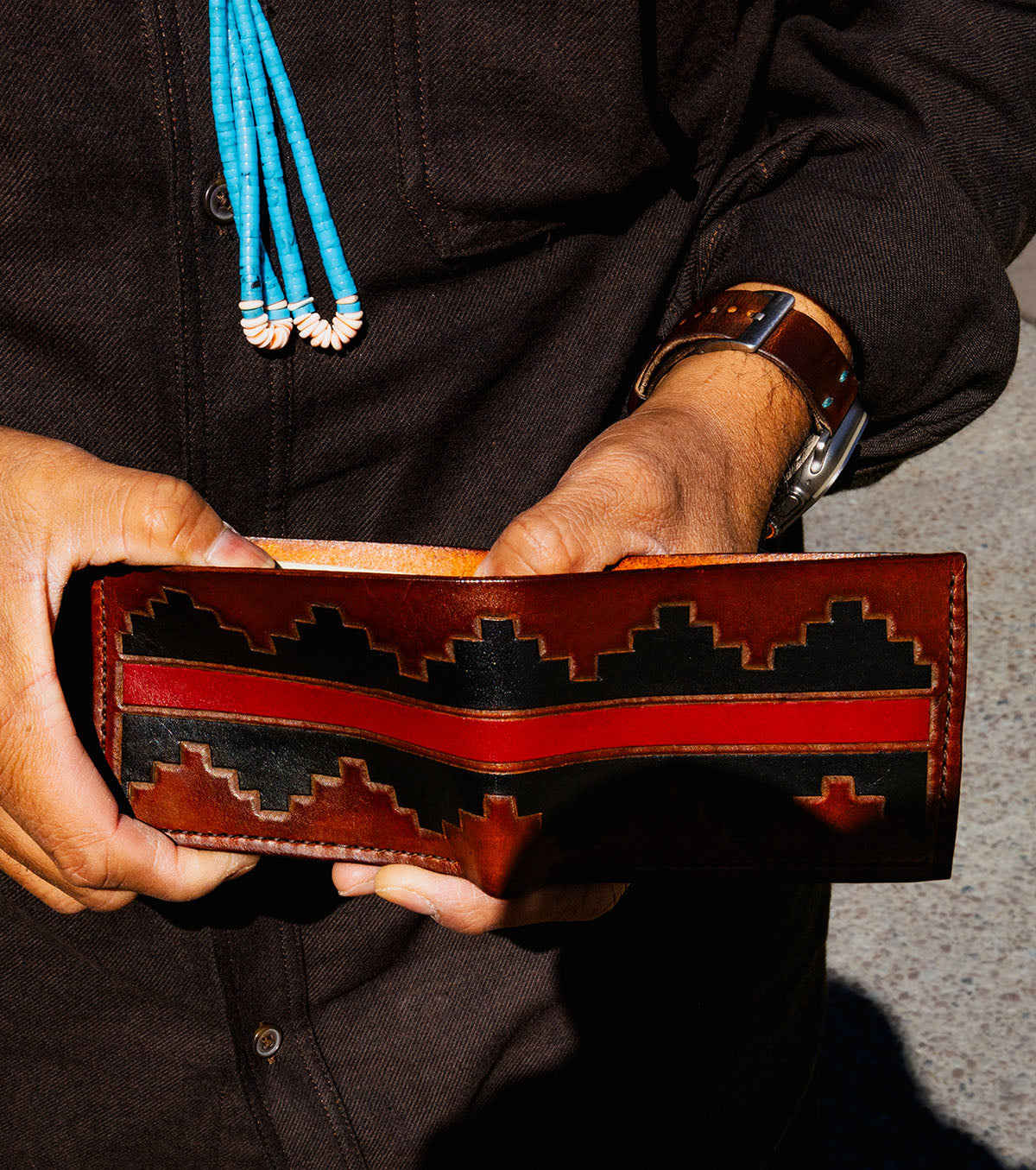A close-up of a person holding a brown leather bi-fold wallet with a black and red geometric pattern. The person is wearing a dark brown shirt, a turquoise and white beaded necklace, and a brown leather watch.